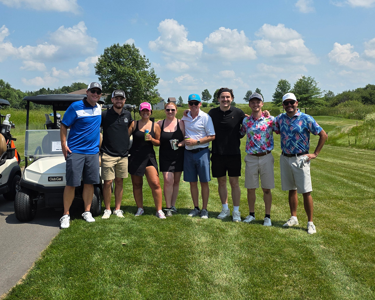 8 adults in summer golf attire pose for a photo on a golf course next to a golf cart. The sky is bright blue with a few fluffy clouds and it's a sunny day. All but one of the adults have hats and/or sunglasses on.
